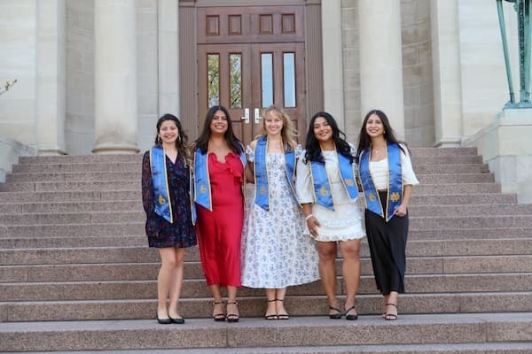 Six women wearing graduation stoles stand in a line on the steps of Bond Hall at the University of Notre Dame.